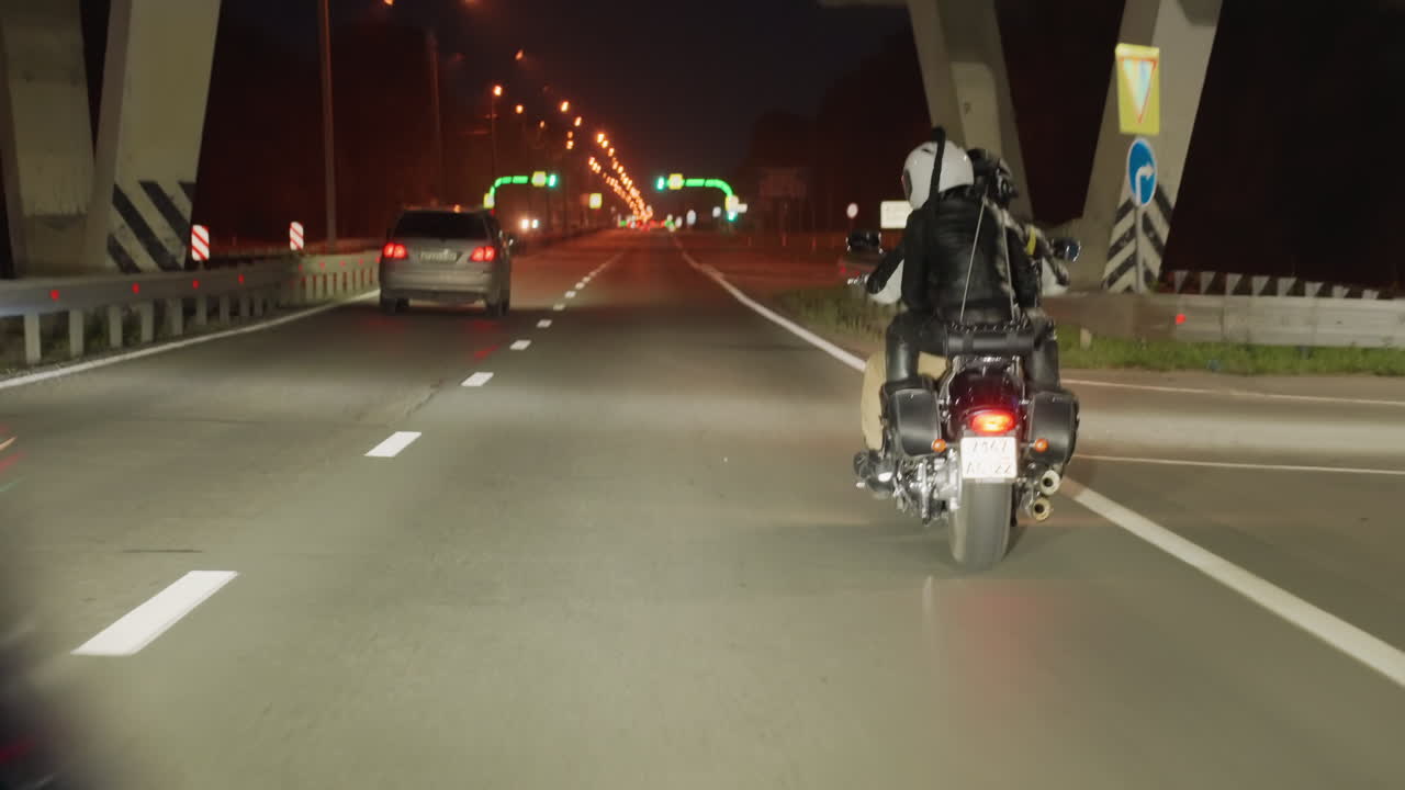 Back view of motorcycle rider with passenger traveling highway at night, riding past bridge structure with cars nearby, heading toward illuminated city road lined with bright streetlights