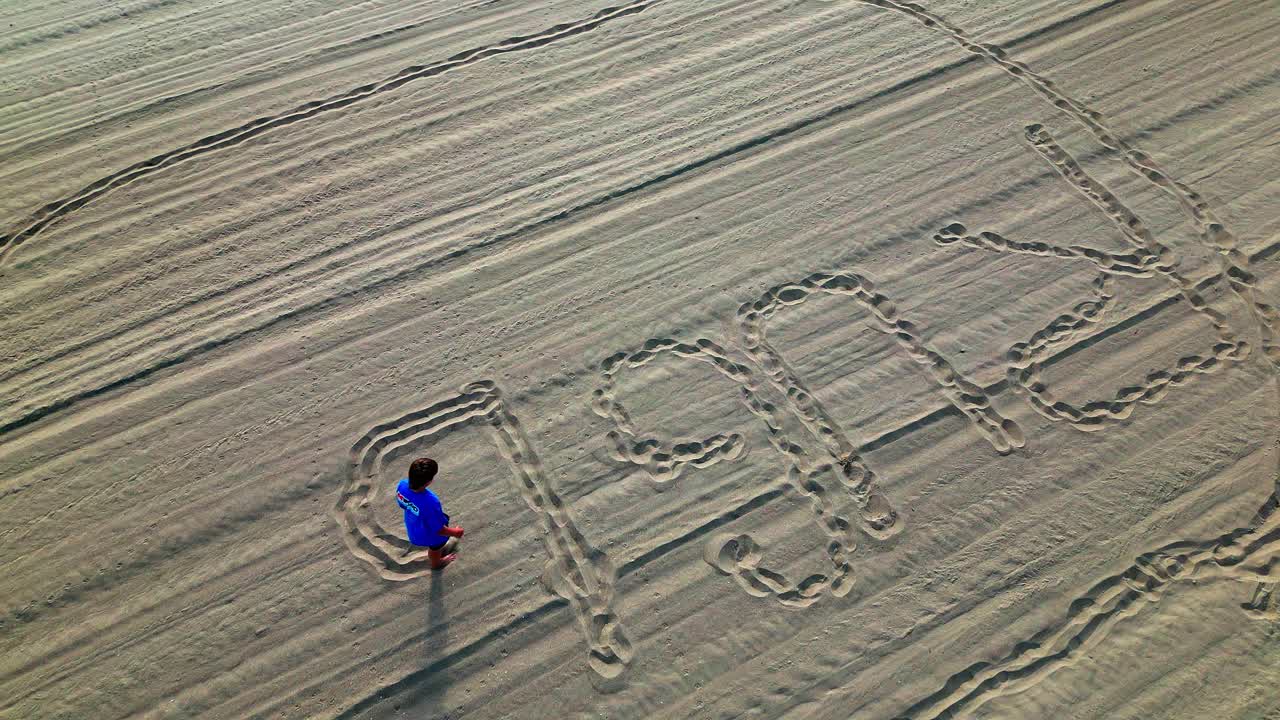 Playful Boy writes name with Bare Feet in Sandy Beach. High angle shot