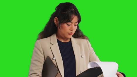 Close-Up-Studio-Portrait-Of-Female-Teacher-Or-Businesswoman-Standing-Against-Green-Screen-Looking-At-Clipboard