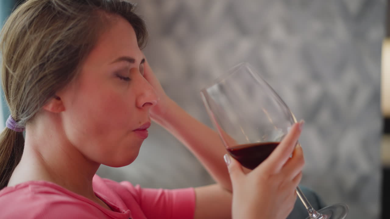 Close up of girl in pink shirt cooling off while drinking red wine with left hand resting on head seated indoors with soft blurred background