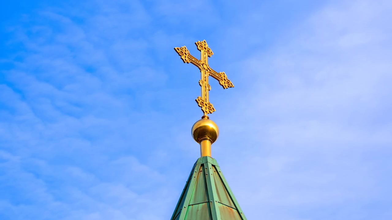 Close view of a gilded Orthodox cross mounted on an orb atop a green copper conical spire, centered against a pale blue cloudy sky. Natural daylight