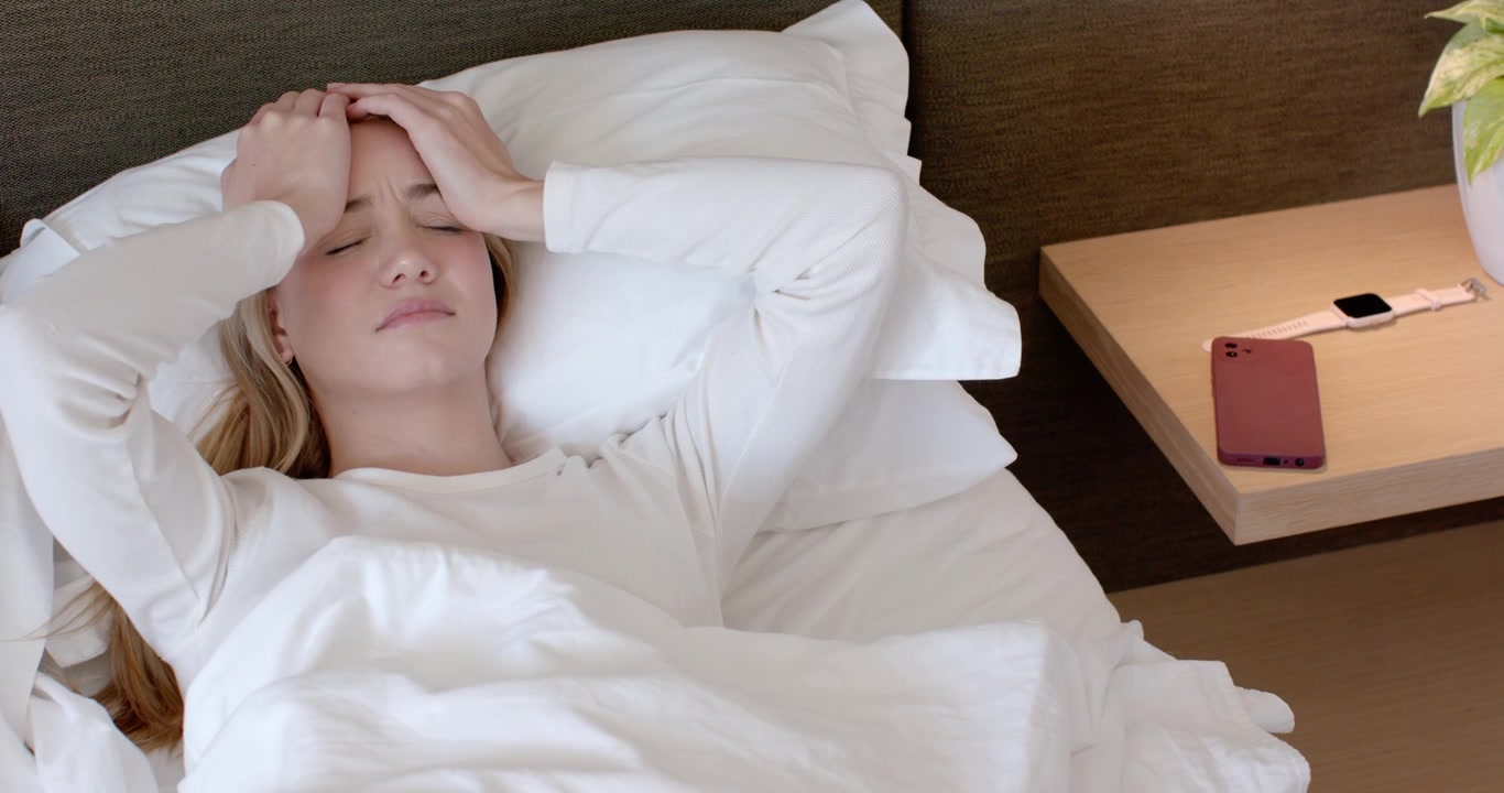 Female in 20s waking from sleep, pressing brow, opening eyes, looking toward bedside to check phone