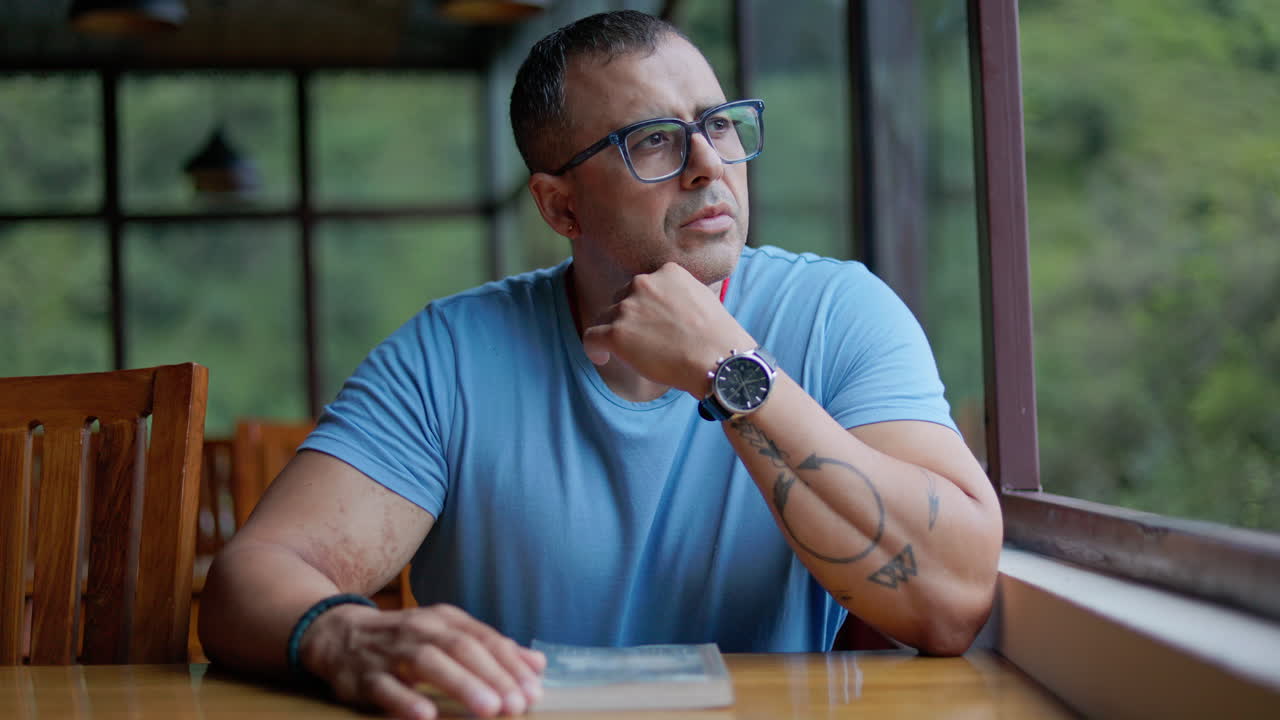 Man in casual outfit sits at wooden table near window with a book, reflecting quietly while looking outside