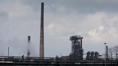 Industrial-plant-with-smokestacks,-steam-clouds-billowing-against-a-cloudy-sky,-evoking-a-moody-atmosphere