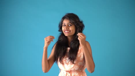 Ein-Junges-Indisches-Mädchen-In-Orangefarbenem-Kittel,-Das-Lächelt-Und-Auf-Der-Hand-Klopft-Und-Auf-Einem-Isolierten-Blauen-Hintergrund-Steht