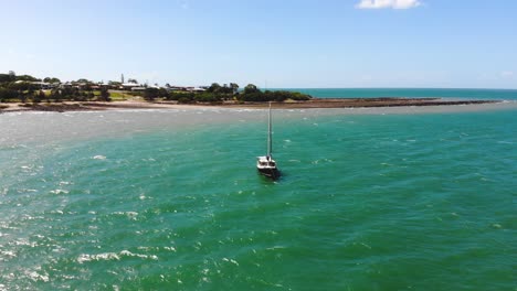 Circling-around-a-small-sailing-boat-that-floats-in-the-tropical,-demin-blue-and-emerald-green-water-just-shy-of-the-shoreline-whilst-a-blue-sky-with-medium-sized,-fluffy-white-clouds-float-above