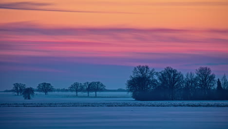 Time-lapse-of-a-snowy-fields-and-bare-trees-against-a-colourful-sky