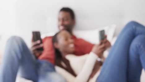 Happy-african-american-couple-lying-on-bed-talking-and-using-smartphones,-in-slow-motion