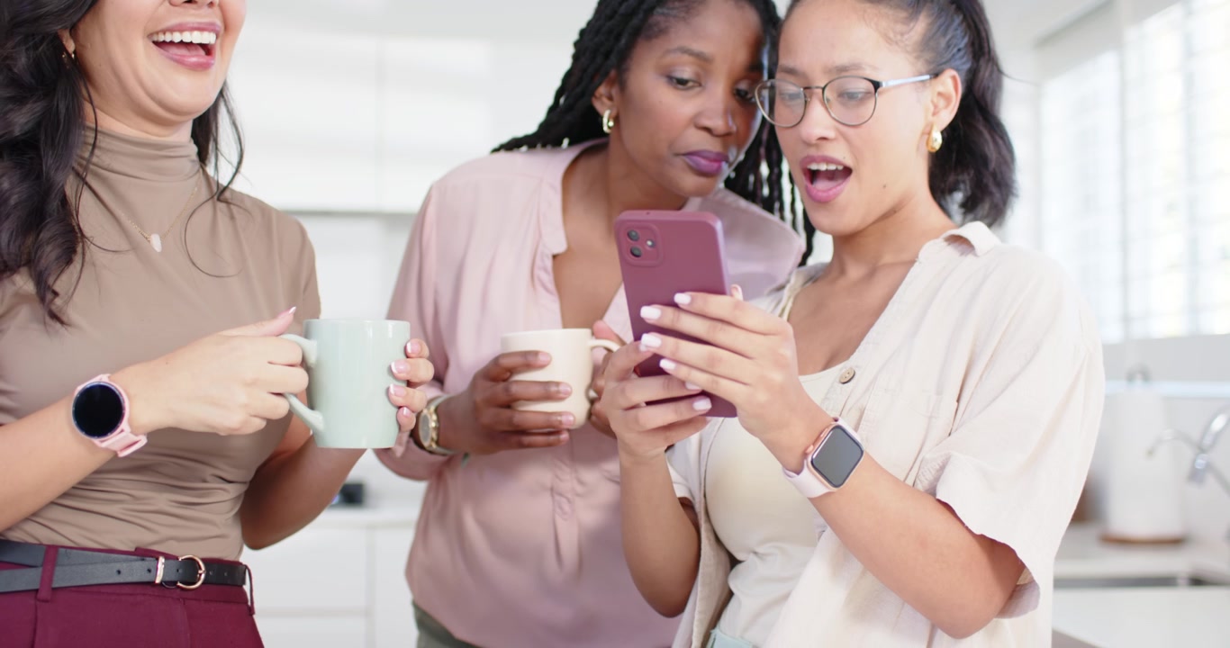 Diverse female friends leaning in white kitchen, right-hand woman showing pink phone, holding mugs