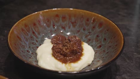 chefs makes plating of meat and mashed potato dish, adding bits of green veggies nn top