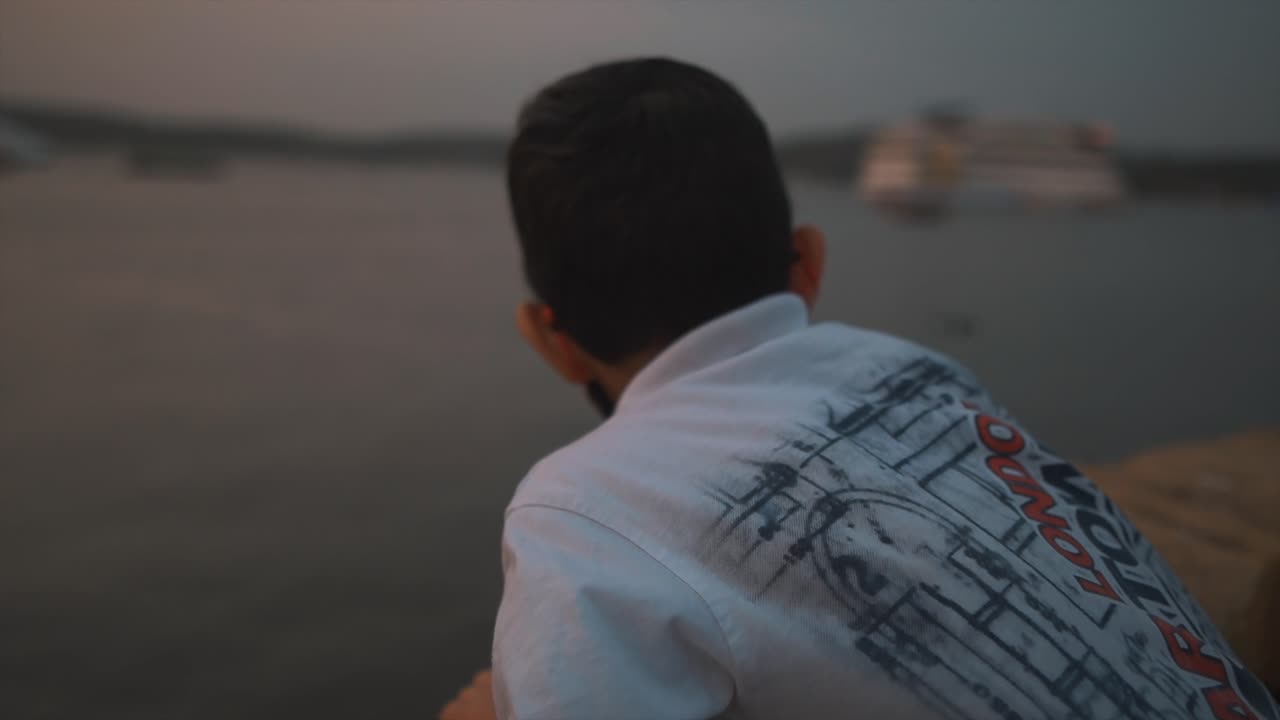 일몰에 마하다이 강을 바라보는 열광적인 어린 소년의 클로즈업, 아름다운 저녁, 고아, 인도의 바다에 정박된 먼 배와 선박을 바라보는 소년