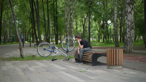 young boy in black top sits thoughtfully on park bench, resting chin on his hand, his bicycle is upside down next to him, and another person walks by in the blurred background