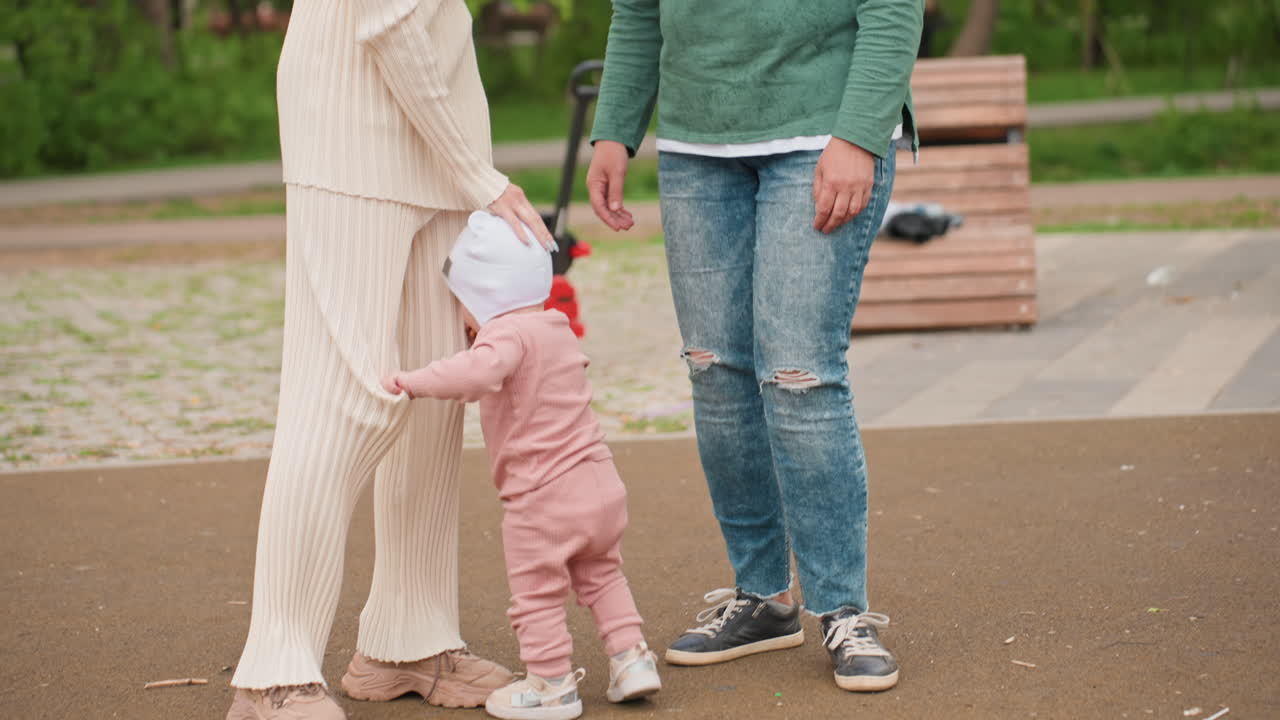 Women Walking Arm In Arm In Green Park, Sharing Quiet Laughter While Toddler Explores Nearby Tire, Accessible Playground In Background With Bench And Trees, Warm Supportive Moment
