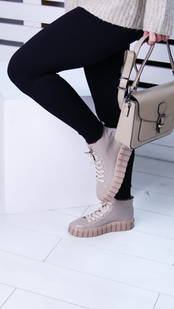 Woman in black tight pants, warm sweater and boots standing in the room. Lady with beige bag demonstrating sneakers. White backdrop. Vertical video