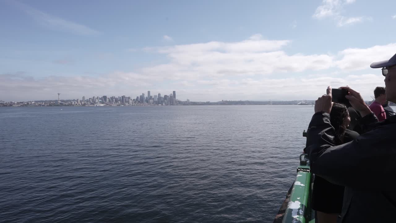 A tourist captures the Seattle skyline with a cellphone while riding a ferry on a clear day, enjoying views of the city and Elliott Bay.