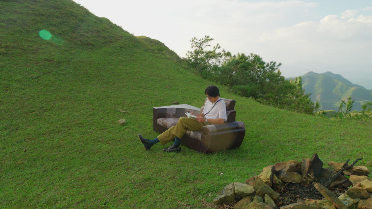 A man reads a book on a couch on a grassy hillside with a mountain view