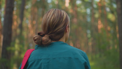 vista trasera de una mujer caminando por un pacífico bosque iluminado por el sol con una mochila roja colgada sobre su hombro, el cabello cuidadosamente atado hacia atrás, la luz del sol se filtra a través de árboles altos