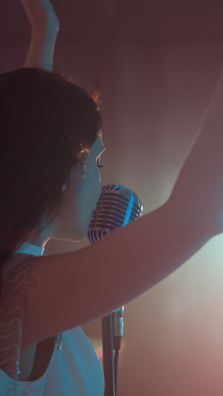 색 드레스를 입은 가수가 빈티지 마이크를 들고 연주하고 있으며, 그녀의 손을 우아하게 움직이고, 배경에는 부드럽고 반이는 불빛이 있습니다.