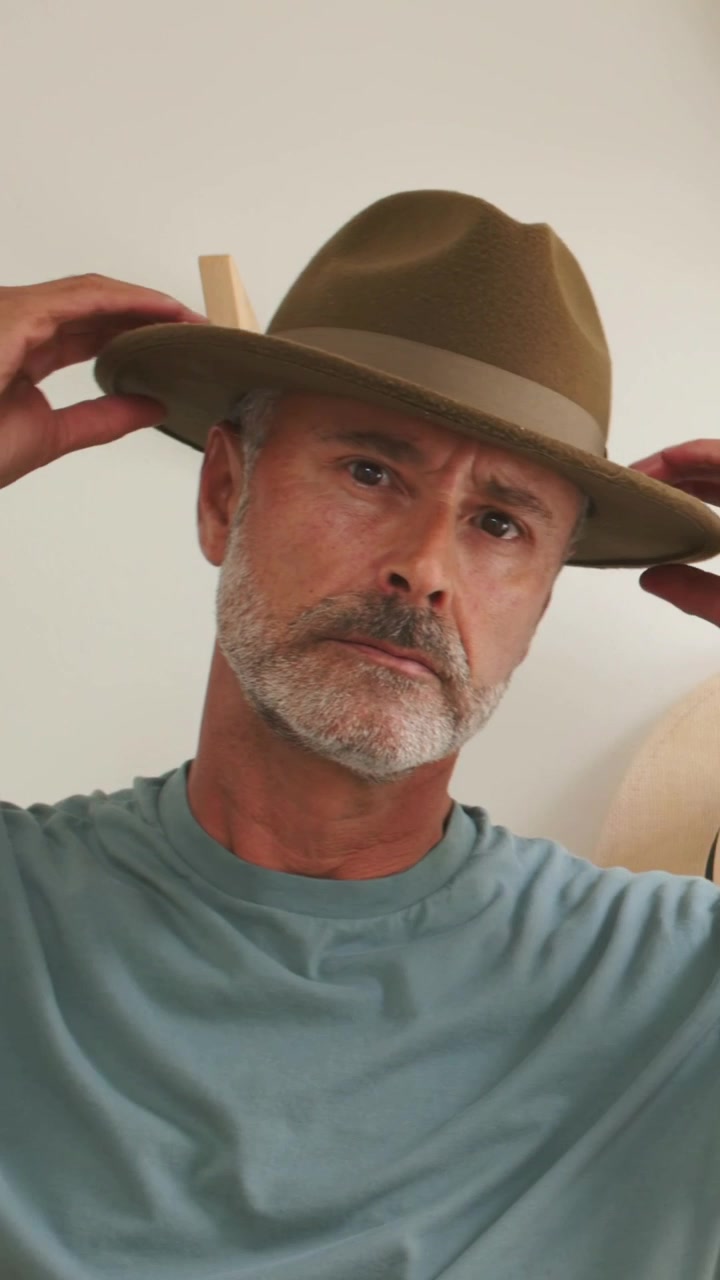 Video vertical: Hombre con camisa verde azulado levantándose el sombrero, ajustándose un fedora marrón cerca de una pared clara, comprobando el ajuste.