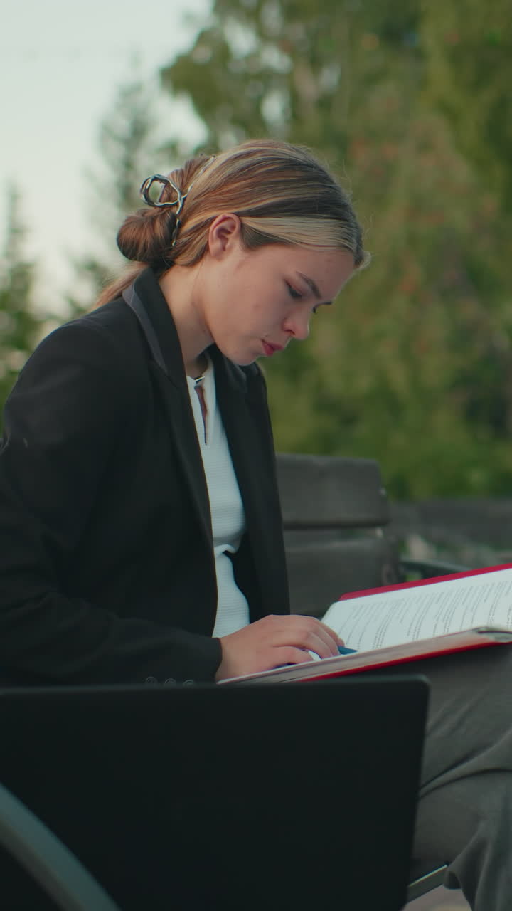 White lady carefully going through her book seated alone outdoors on wooden bench with laptop nearby in quiet urban setting surrounded by empty benches trees and pole in background