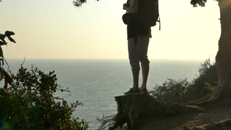 young man wearing a large travel backpack standing in silhouette on a tree stump looking out over a pacific ocean cliff during a golden sunset in santa barbara, california