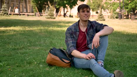 young handsome man is sitting on grass in park, listening music with earphones, relax concept