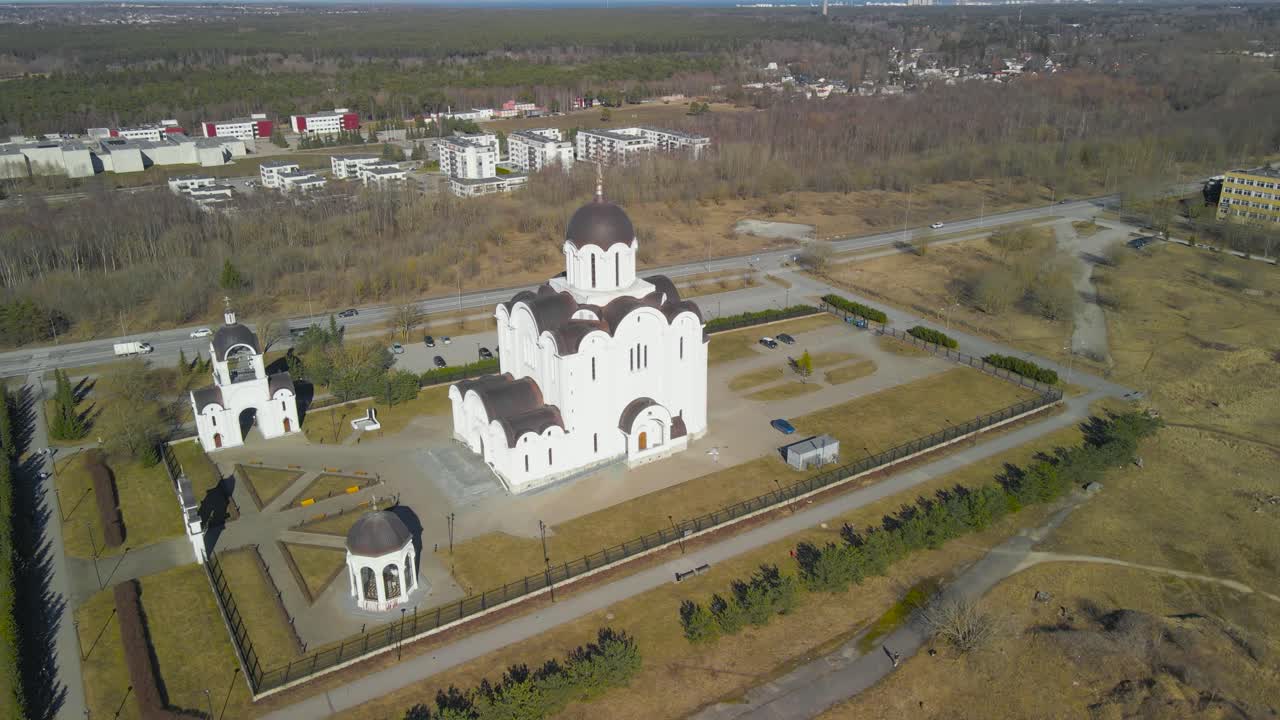 Aerial drone footage circling around and orbiting a large white russian church in Lasnamäe Estonia during a sunny spring day with large empty fields that are brown and green are surrounding it.
