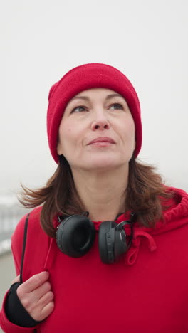 close-up of woman with headphones around neck walking along interlocked pathway with bag over shoulder and smile near iron railing overlooking blurred cityscape and light poles