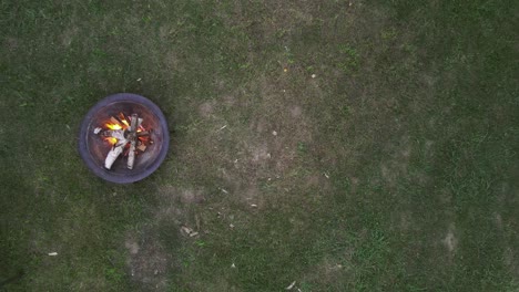 Aerial-birdseye-view-of-a-fire-pit-on-the-grass-with-people-walking-buy-and-someone-putting-wood-into-the-fire