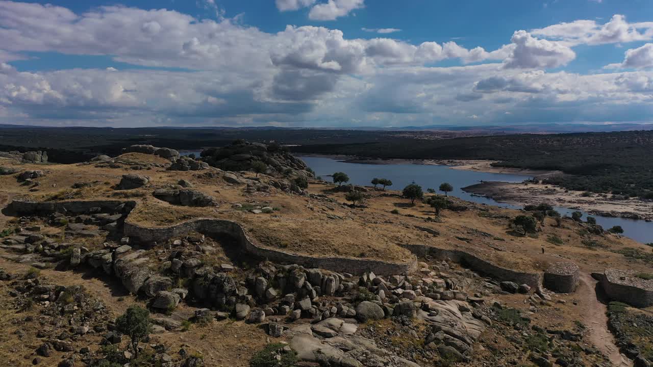 vuelo lateral en un fuerte celta vetton de la edad de hierro viendo la entrada a la ciudad y su extensa pared de piedra reconstruida al final vemos la presa del embalse en avila españa