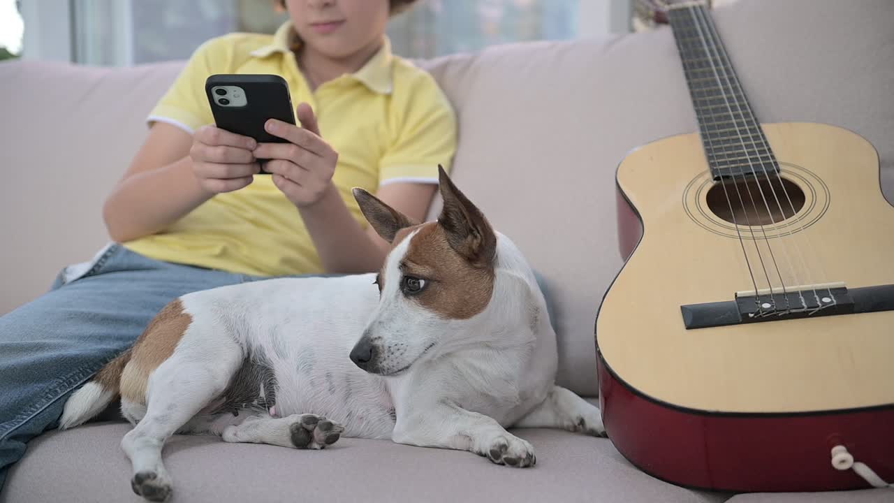 기타 근처에 누워 있는 개 옆에 스마트폰을 사용하여 소파에 앉아 있는 금발 소년