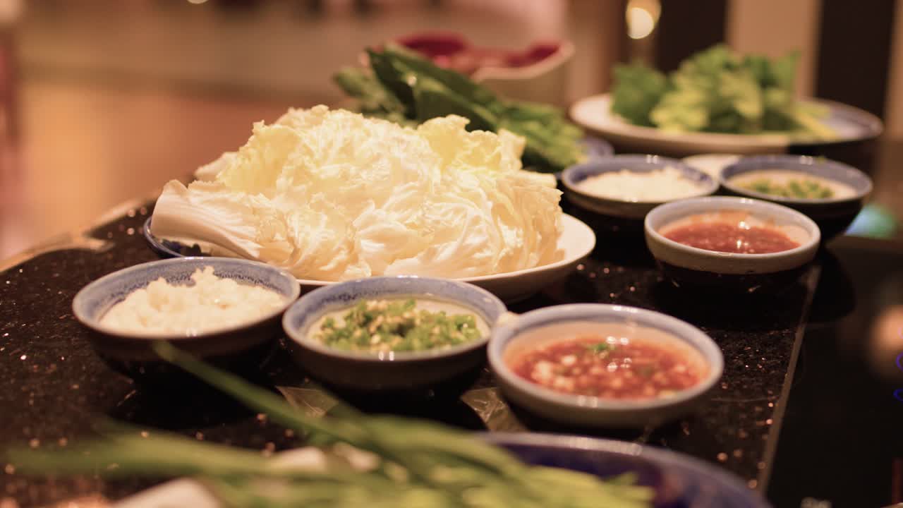 Assorted vegetables, sauces, and rice arranged for hotpot, warm lighting, slight camera pan right