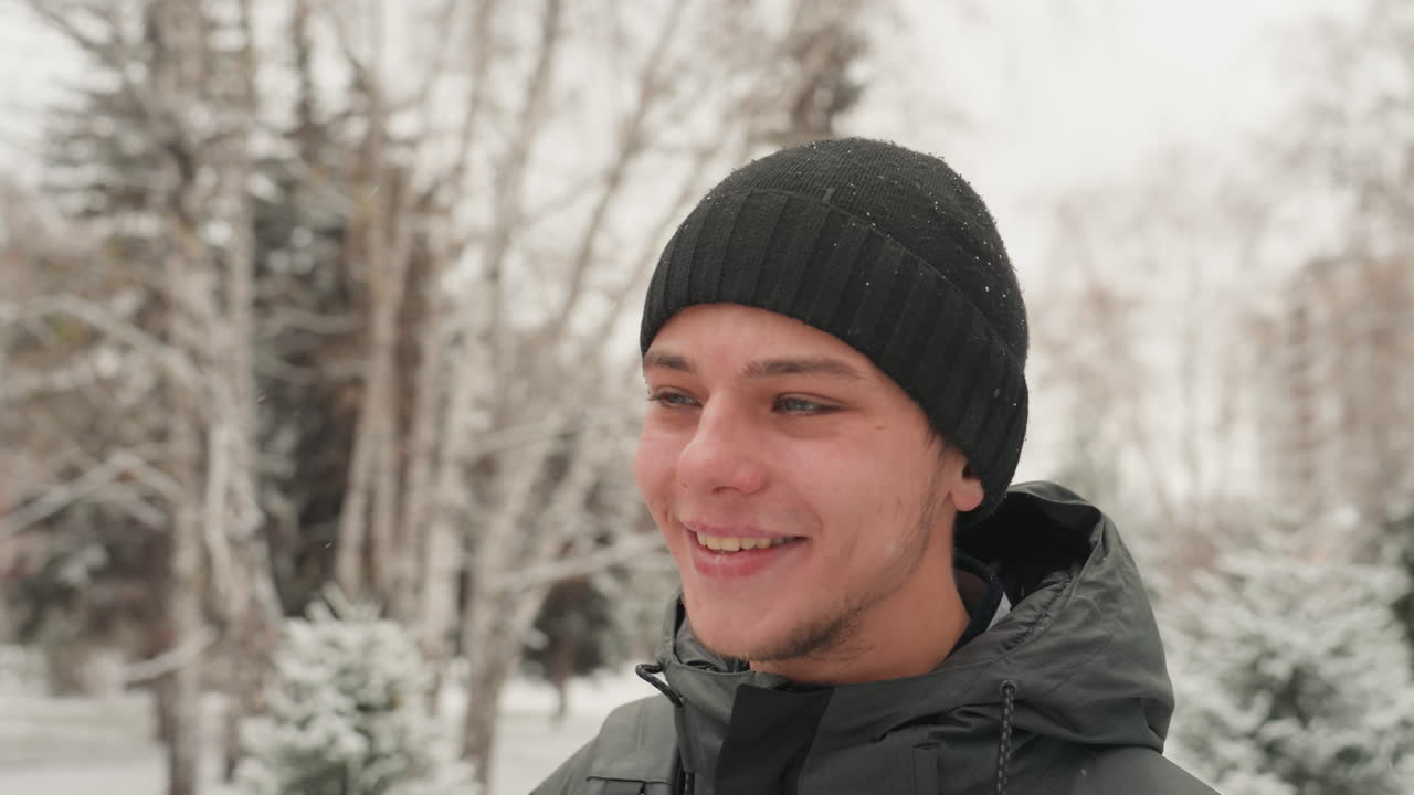 Młody biały mężczyzna śmiejący się w śniegu, uśmiechający się szeroko z ożywioną ekspresją, w czarnej czapce i zimowej kurtce, z lekkimi płatkami śniegu na rzęsach, w naturalnym, swobodnym nastroju, na tle wiecznie zielonych drzew, radosny.