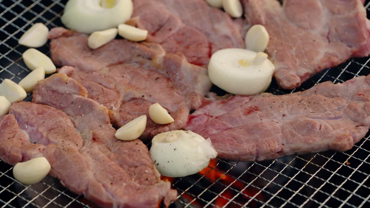 An extreme close-up shot captures seasoned pork slices, garlic cloves, and onions sizzling on a hot barbecue grill over charcoal flames, a popular Korean-style outdoor meal
