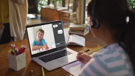 Schoolgirl-using-laptop-for-online-lesson-at-home,-with-boy-talking-and-web-chat-on-screen