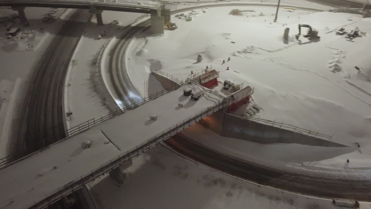 Multiple snow plows work to clear a snow-covered highway during nighttime traffic, surrounded by glowing streetlights, active vehicles, and fresh snowfall in a winter urban setting.