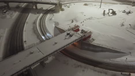 multiple snow plows work to clear a snow-covered highway during nighttime traffic, surrounded by glowing streetlights, active vehicles, and fresh snowfall in a winter urban setting.