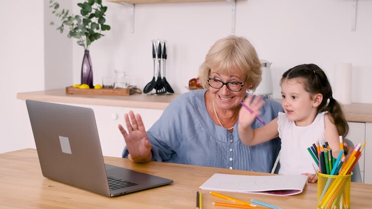 Little Girl Drawing Sitting At Table In Kitchen With Her Happy Grandmother While Having A Video Call On Modern Laptop
