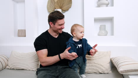 father is holding a baby by hands standing on the sofa. dad is smiling to his son trying to dance.