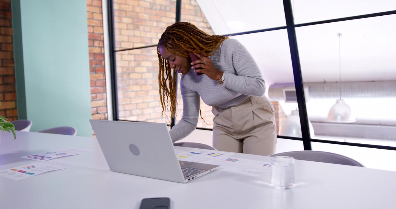 Woman taking phone call while leaning over table, using laptop and reviewing charts for work