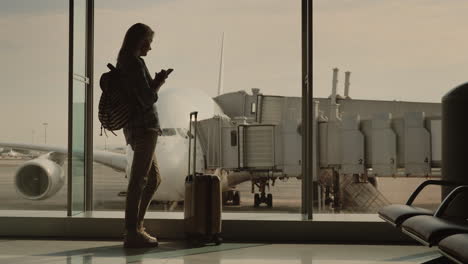 a young woman with a backpack and a suitcase on wheels looks out the window at the plane and uses a