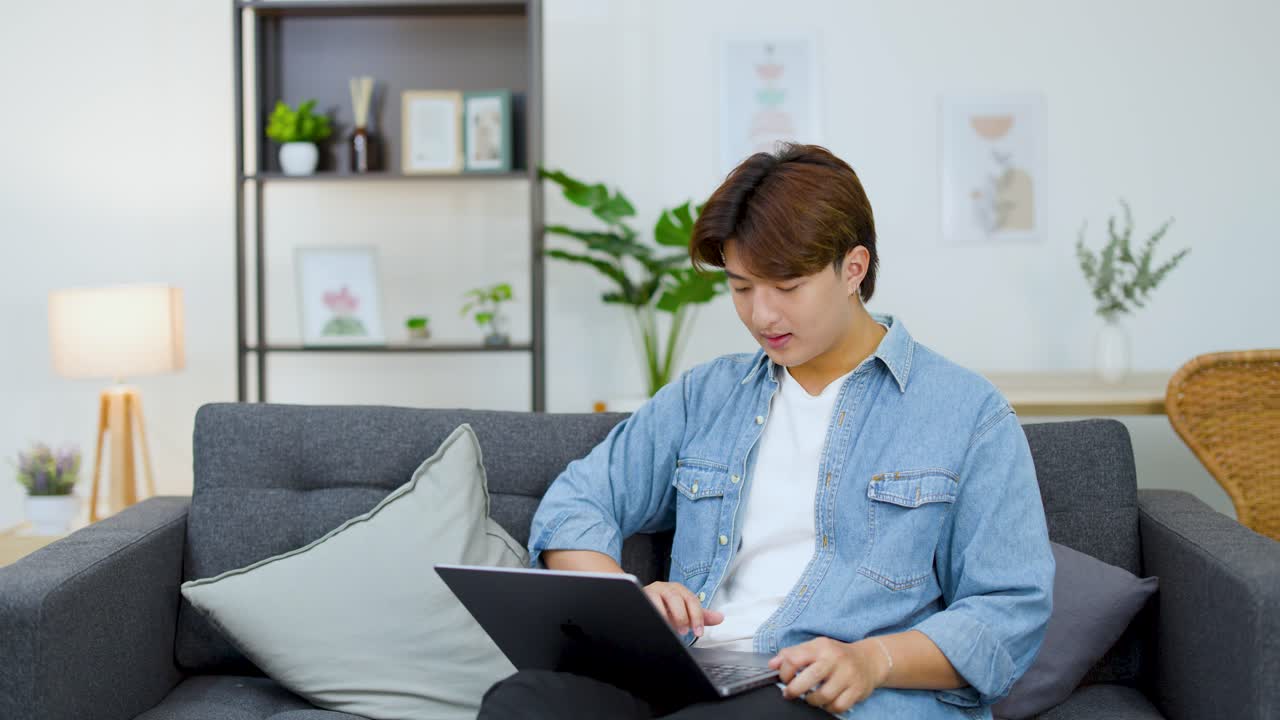 若いアジア人男性がソファに座り、ノートパソコンとクレジットカードを使って買い物している様子。