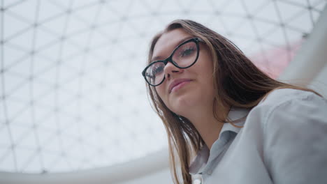 up de la dama en la escalera mecánica con gafas mirando contemplativa, enmarcado contra un techo interior moderno, reflejando la expresión reflexiva en un entorno urbano