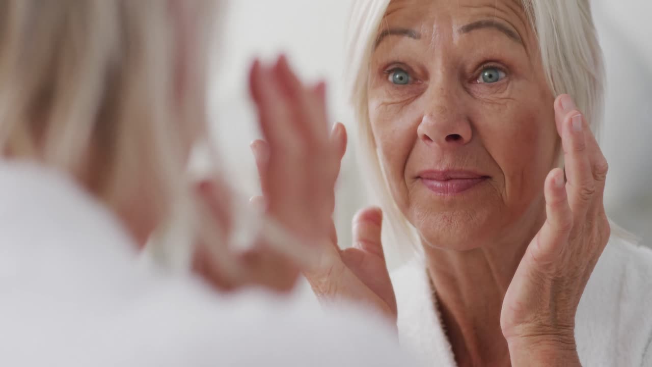 Happy senior caucasian woman looking at mirror in bathroom and touching her face
