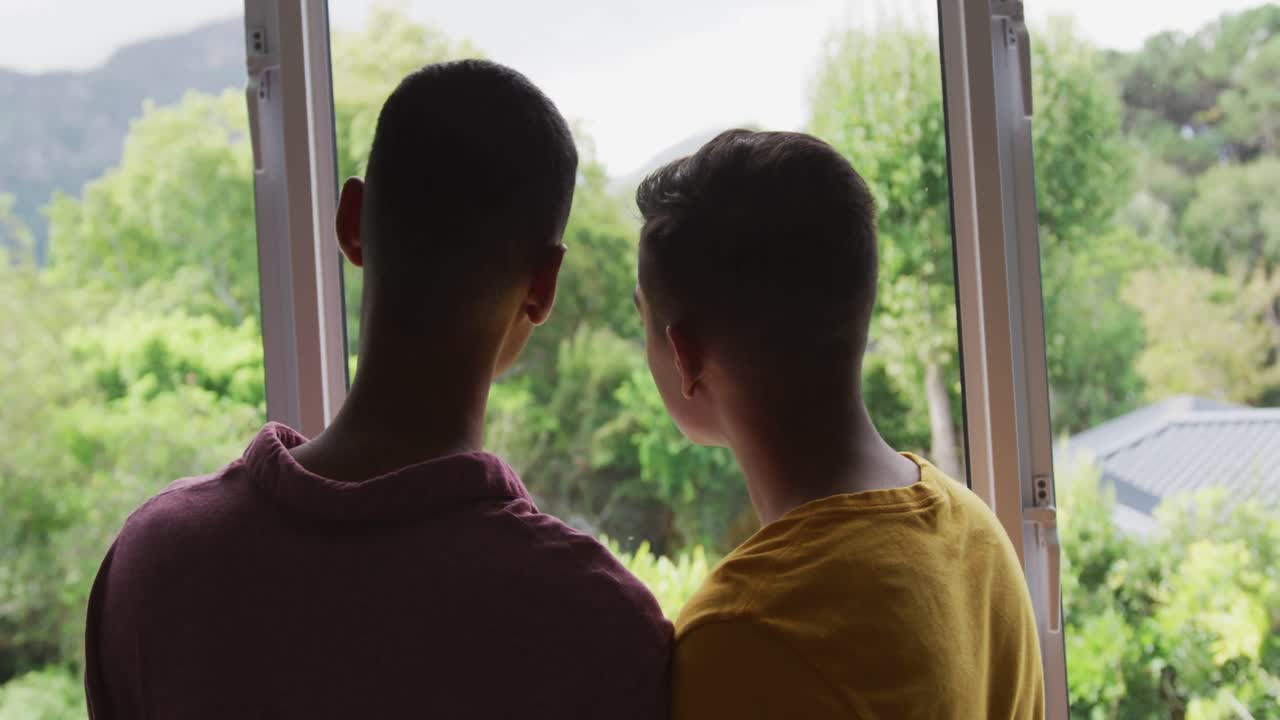 Rear view of mixed race gay male couple standing looking out of window leaning heads together