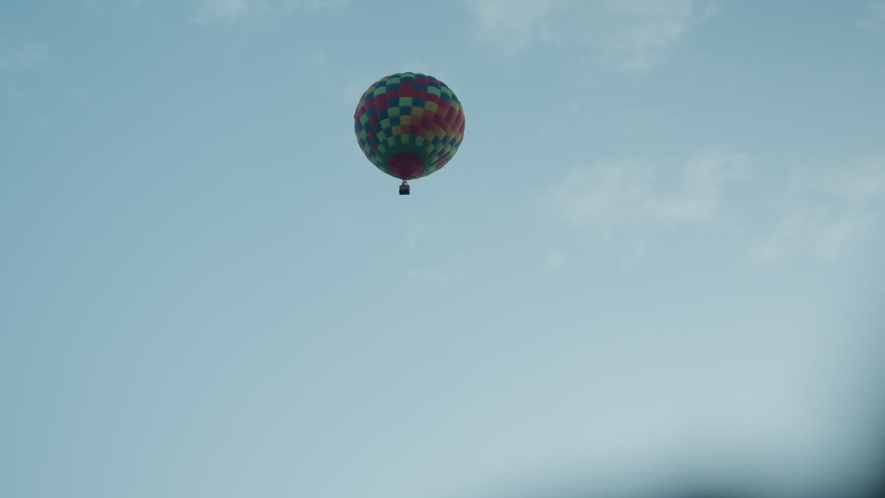 distant bright patchwork hot air balloon ascending above open green field with blurred dark foreground shape against clear blue sky at dawn evoking sense of scale adventure
