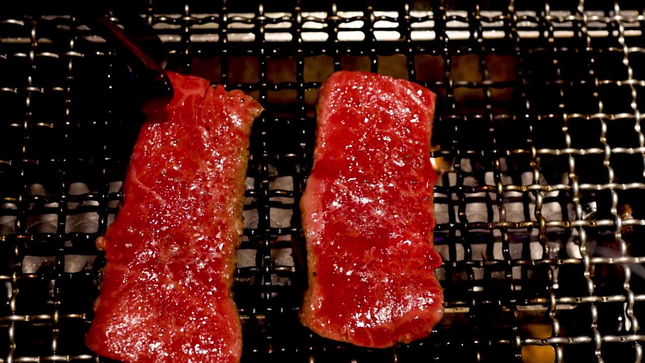Two steaks sizzling on a grill, with tongs flipping them for even cooking.
