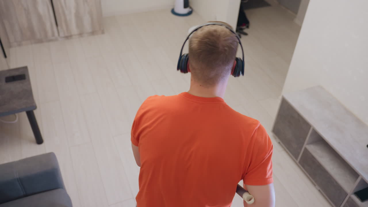 Overhead perspective of young man in orange shirt wearing headphones joyfully dancing while mopping tiled floor in modern living room, expressing energy and enjoyment through cleaning task