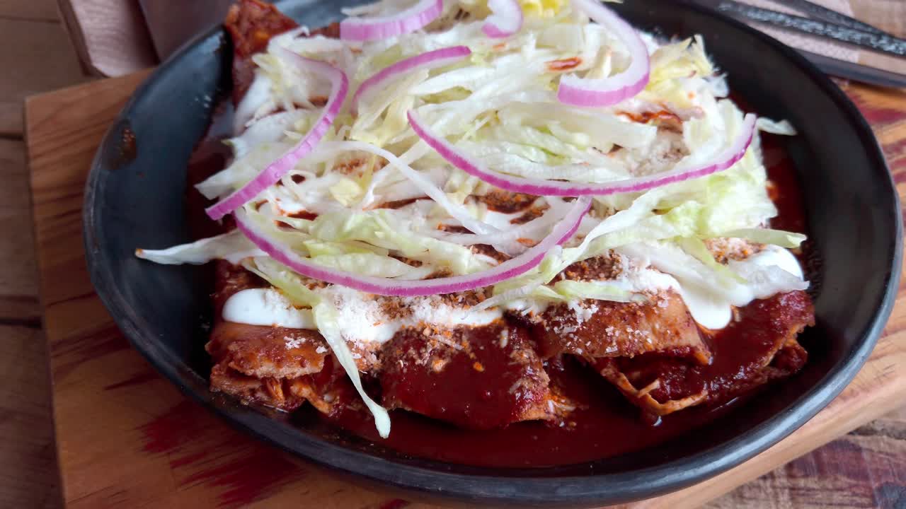 Enchiladas topped with sour cream, shredded lettuce, red onion rings, and crumbled cheese on a dark plate. Close up rotating view