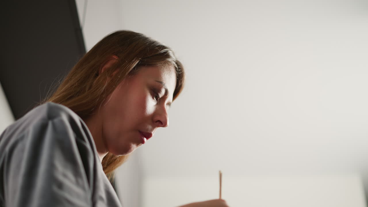 Side view of hungry girl eating dinner while standing in bright kitchen, soft light reflecting off her face, focused on meal, dressed in casual grey shirt, modern minimalistic background